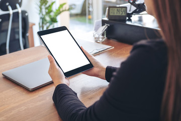 Mockup image of a woman holding and using black tablet pc with blank white desktop screen with notebook on wooden table in office