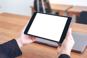 Mockup image of hands holding and using black tablet pc with blank white desktop screen with notebook on wooden table in office