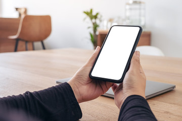 Mockup image of hands holding black mobile phone with blank white screen with laptop on wooden table