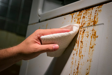 Hand cleaning baked on kitchen grime on side of oven appliance, using paper towel and cleaner.