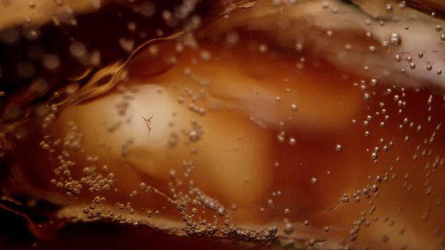 Bubbles fizzing in cola drink around ice in macro view in glass.