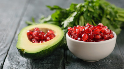 Grains of ripe pomegranate in a white bowl and in a half of avocado on a dark table.