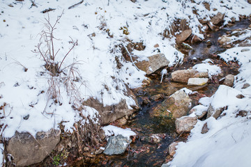 Meltwater stream running through the stones and surrounded by brown melting snow and dry grass at mountains.