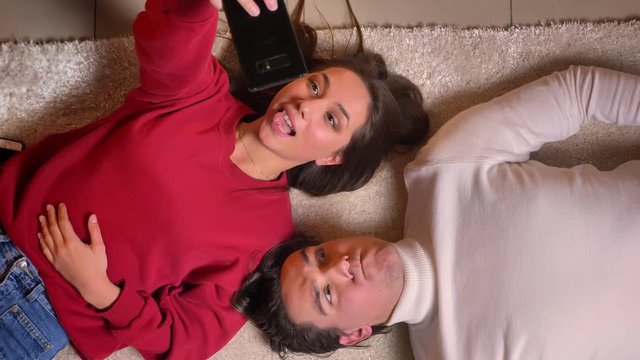 Top shot of young couple lying head to head on carpet and making funny selfie-photos using smartphone in cosy atmosphere.