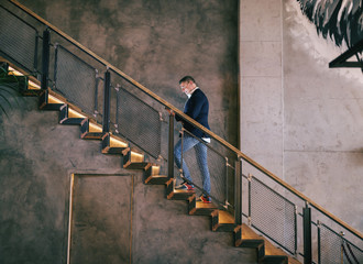 Middle-aged man dressed smart casual climbing the stairs and using tablet for video call. Full...