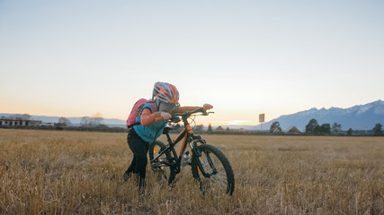 Obraz premium One caucasian children walk with bike in wheat field. Little girl walking black orange cycle on background of beautiful snowy mountains. Biker stand with backpack and helmet. Mountain bike hardtail.