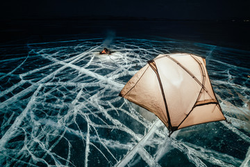 Fire on ice at night. Campground on ice. Tent stands next to bonfire. Lake Baikal. Nearby there is car. Shelter tent and ice are illuminated from the inside. Beautiful bonfire on real cracked ice © ivandanru