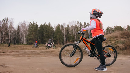 Obraz premium One caucasian children rides bike road track in dirt park. Girl riding black orange cycle in racetrack. Kid goes do bicycle sports. Biker motion ride with backpack and helmet. Mountain bike hardtail.