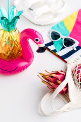 Summer vibes concept with colorful travel fashion items, sunglasses, scarf, pink dragon fruit, flamingo inflatable drink holder, pineapple straw tumbler bottle, net bag and sandal on a white backgroun