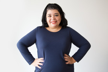 Smiling overweight Hispanic woman posing with hands on hips. Successful adult woman standing with...