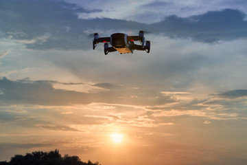 Fototapeta na wymiar A small smart quadcopter hovering in the air against setting sun and colorful evening sky. Drone flying in the nature, illuminated by setting sun.
