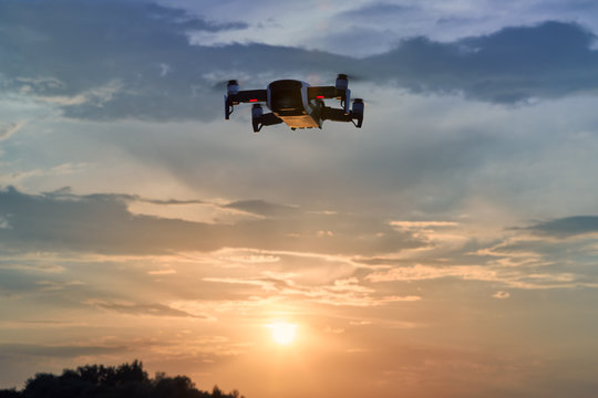 A small smart quadcopter hovering in the air against setting sun and colorful evening sky. Drone flying in the nature, illuminated by setting sun.