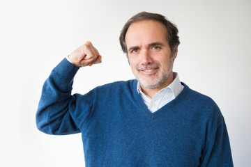 Successful strong man showing muscular arm and looking at camera. Handsome positive man in blue sweater holding fist up. Isolated on white. Success concept