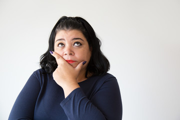 Thoughtful overweight woman holding chin and looking up, studio portrait. Pensive Latin woman in...