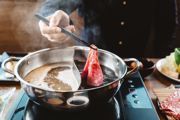 Man holding rare slice Wagyu A5 beef into shabu hot pot shoyu soup base by chopsticks with steam.