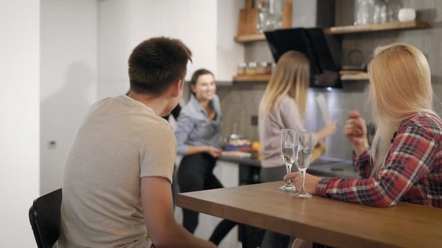 five young people are resting on small home party in vacations, pair is drinking wine and they friends are cooking
