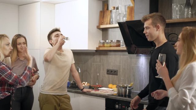 cheerful teenage boys and girls are having fun on a kitchen in apartment in evening, drinking wine