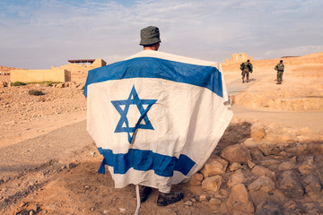 Israeli military infantry stands in middle of the desert holding an Israeli flag with the star of...