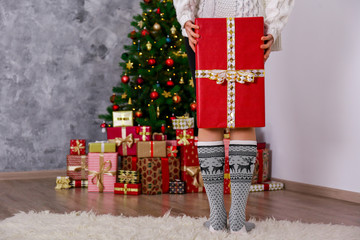 Cropped shot of female legs standing, wearing mid-calf socks with reindeer print and ornament, Decorated christmas tree on background. Copy space, close up.