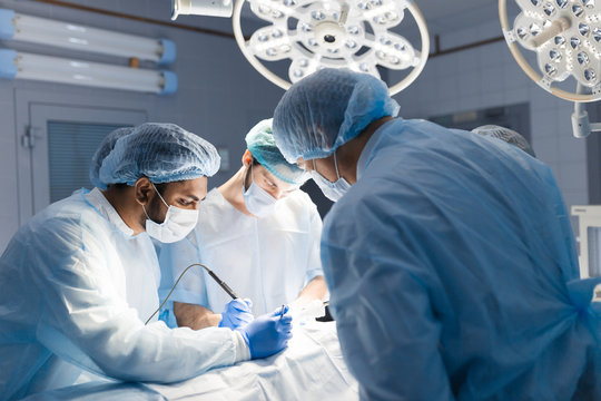 Surgeon doctor operating using electric scalpel wearing blue surgical mask and surgical cap in surgery room with his team surgeons operating