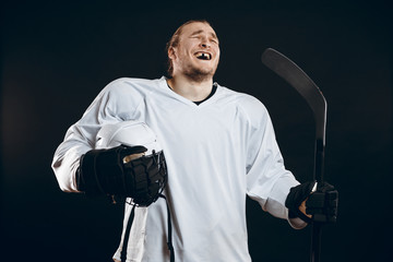 Happy Handsome hockey player with one broken front tooth laughing at camera, standing with stick in white uniform, isolated on black