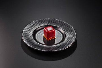 Miniature cake in red glaze has the shape of a cube on a black plate.