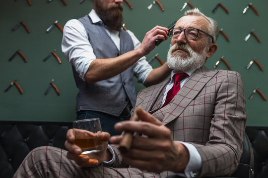 Portrait of relaxed grey haired businessman with sigar and alcoholic beverage having his hair trimmed in a barber shop, low angle. Concept of grooming
