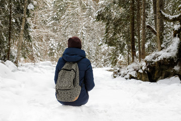 Fototapeta na wymiar Girl with backpack sitting in a snow-covered coniferous forest. Winter cloudy day.