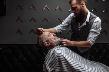 Caucasin old man getting his beard shaved by barber visiting hairtician at shaving saloon.