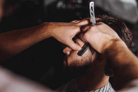 Skillful professional barber at work. The hands of young barber shaving with a razor handsome brunette man in barbershop, close up.
