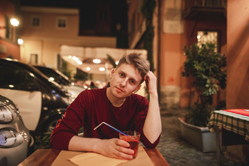 Handsome young man sits in the evening on the terrace of a restaurant with a glass of alcohol in his hands and looks at the camera. Evening holidays in the cozy restaurant on the terrace.