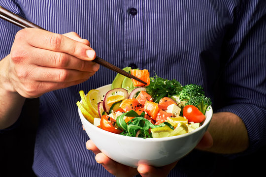 man in a shirt holds poke bowl with salmon, avocado, cucumbers, arugula, broccoli, rice, carrots, cheese and chuha with chopsticks