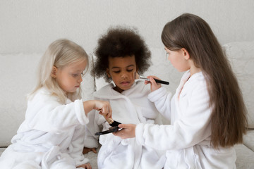 Little girls play with makeup while sitting on the couch. Girls of different nationalities paint their faces with makeup poGirls of different nationalities paint their faces with makeup powder cheeks.
