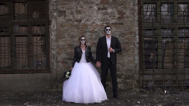 A horrible couple in a wedding dress and costume with makeup in the form of a skeleton stand against an old brick wall in an old abandoned house. The concept of Halloween. Halloween face art.