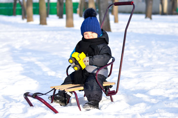 Fototapeta na wymiar Cute little boy sitting in sleigh in winter park all around the snow