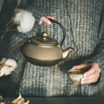 Tea ceremony. Young female in grey warm knitted winter sweater pouring green tea from golden iron pot into japanese ceramic cup in kitchen, square crop. Cold winter morning at home