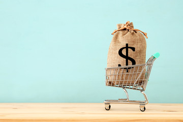 shopping cart with bag full of money with dollar sign over blue wooden background © tomertu