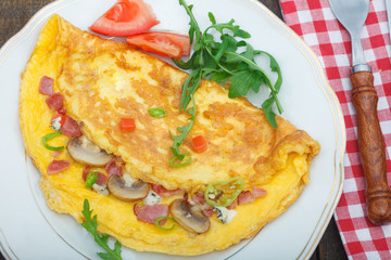 Omelette with ham, mushrooms and tomatoes. Homemade omelette with rucola green salad on a white plate and vintage wooden table.Top view