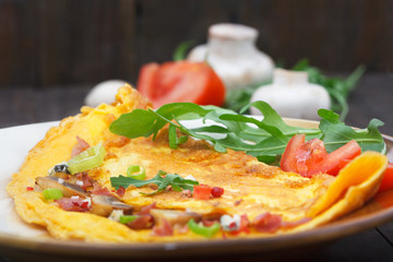 Omelette with ham, mushrooms and tomatoes. Homemade omelette with rucola green salad on a plate and vintage wooden table.