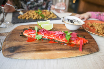 sea bass fillet with red sauce, marinated onion, parsley over plant leaf, with slices of lime and bowl with rice, on wooden tray on restaurant table