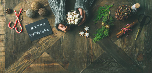 Christmas, New Year preparation background. Flat-lay of greeting card, glittering toy, woman's hands in sweater holding mug of hot chocolate, cinnamon, scissors over wooden table, top view, copy space