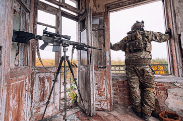 Sniper in a bulletproof vest and helmet looks out the window near a large sniper rifle