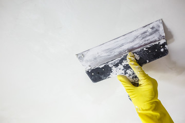 worker's hand in yellow gloves holding putty knife patching a hole with spatula with plaster or...