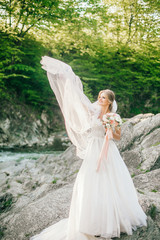 Beautiful luxury young bride in long white wedding dress and veil standing near river with mountains on background