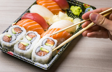 Hand using chopsticks pick Sushi and Sashimi rolls on a black stone slatter. Fresh made Sushi set with salmon, prawns, wasabi and ginger. Traditional Japanese cuisine