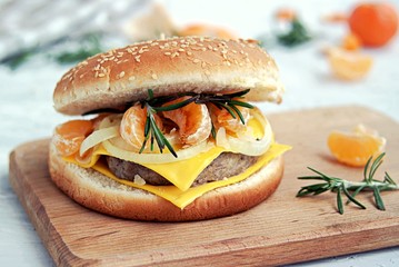 Large homemade burger with juicy meat patty, two slices of cheese, fried tangerines, onions and rosemary on a wooden board.