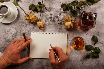 Mock up background with men's hand holding a glass of whiskey, brandy withered flowers of white roses with a clean envelope, notepad and cup of coffee on a gray concrete background. View from above. 