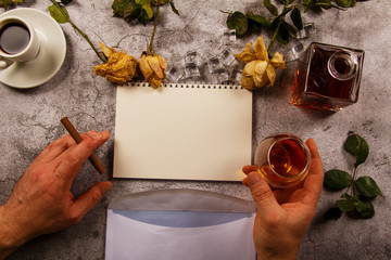 Mock up background with men's hand holding a glass of whiskey, brandy withered flowers of white roses with a clean envelope, notepad and cup of coffee on a gray concrete background. View from above. 