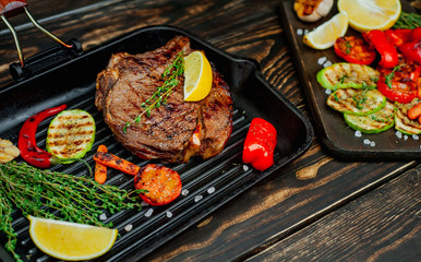 steak in a grill pan, next to chopped vegetables, lemon, red pepper, garlic and seasonings on a dark rustic background