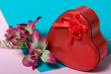 A box in the form of a red heart with a gift. Next is a flower. On a background of multicolored paper.